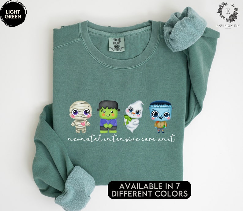 May include: A light green sweatshirt with a design of four cartoon monsters. The monsters are a mummy, a Frankenstein, a ghost, and a zombie. The text 'neonatal intensive care unit' is printed below the monsters. The text 'AVAILABLE IN 7 DIFFERENT COLORS' is printed below the design.