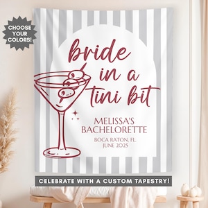 May include: A gray and white striped tapestry with the text "bride in a tini bit" in maroon. The text "Melissa's Bachelorette" is also in maroon, along with the location and date "Boca Raton, FL. June 2025". The tapestry also features a martini glass with olives in it.