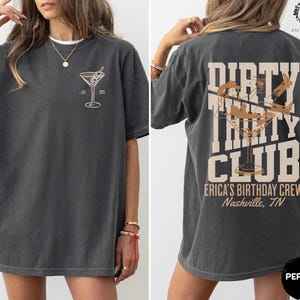 May include: A dark gray t-shirt with a martini glass graphic on the front and the text "Dirty Thirty Club" with a martini glass graphic on the back. The text on the back also includes "Erica's Birthday Crew" and "Nashville, TN".