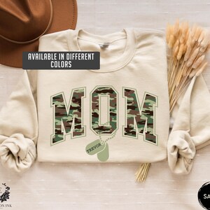 May include: A beige sweatshirt with the word "MOM" in camouflage print. The word "MOM" is in a large, bold font. Below the word "MOM" is a dog tag with the name "TREVOR" on it.