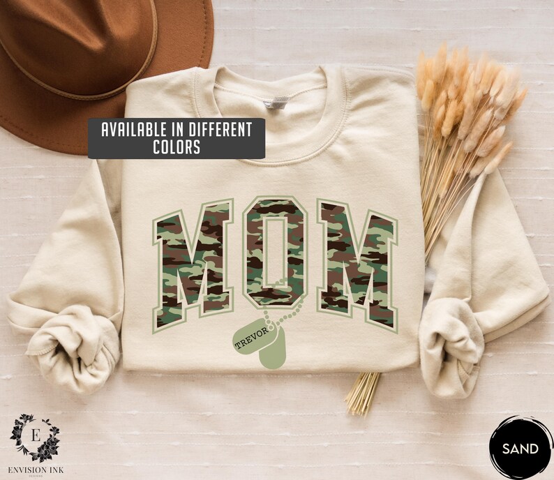 May include: A beige sweatshirt with the word "MOM" in camouflage print. The word "MOM" is in a large, bold font. Below the word "MOM" is a dog tag with the name "TREVOR" on it.