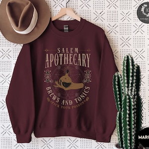 May include: Maroon sweatshirt with a graphic design of a hand holding a potion bottle with the text "Salem Apothecary Brews and Tonics Pick Your Poison" in a vintage style font.
