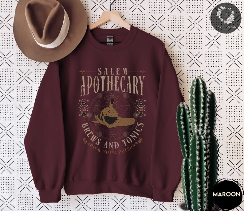 May include: Maroon sweatshirt with a graphic design of a hand holding a potion bottle with the text "Salem Apothecary Brews and Tonics Pick Your Poison" in a vintage style font.