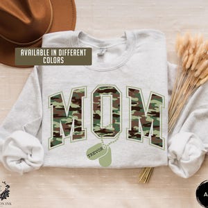 May include: A light gray sweatshirt with a camouflage print of the word "MOM" and a dog tag with the name "TREVOR" hanging from it. The sweatshirt is available in different colors.