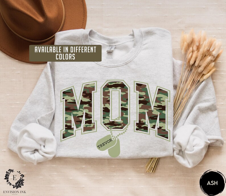 May include: A light gray sweatshirt with a camouflage print of the word "MOM" and a dog tag with the name "TREVOR" hanging from it. The sweatshirt is available in different colors.