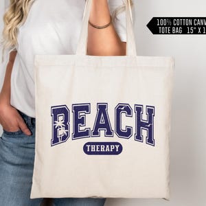 May include: A white canvas tote bag with the text "BEACH THERAPY" in navy blue lettering. The text is in a collegiate font with palm trees and seagulls. The bag is 15 inches by 16 inches.