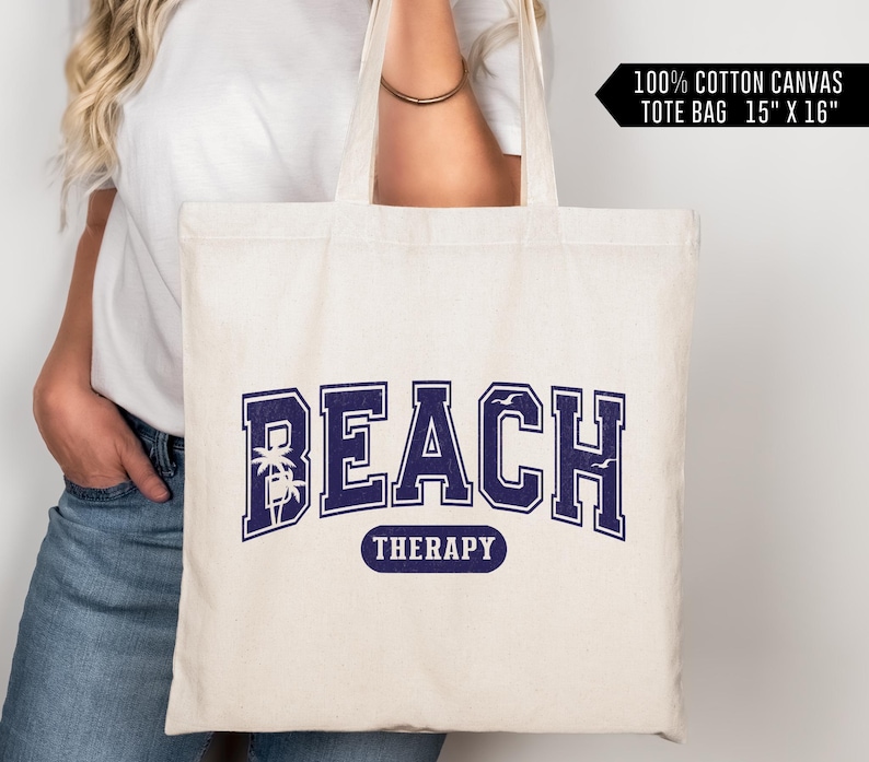 May include: A white canvas tote bag with the text "BEACH THERAPY" in navy blue lettering. The text is in a collegiate font with palm trees and seagulls. The bag is 15 inches by 16 inches.