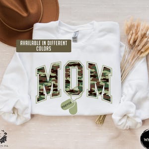 May include: White sweatshirt with a camouflage print of the word "MOM" and a dog tag with the name "TREVOR" on it. The text "AVAILABLE IN DIFFERENT COLORS" is at the top of the image. The word "WHITE" is at the bottom of the image.