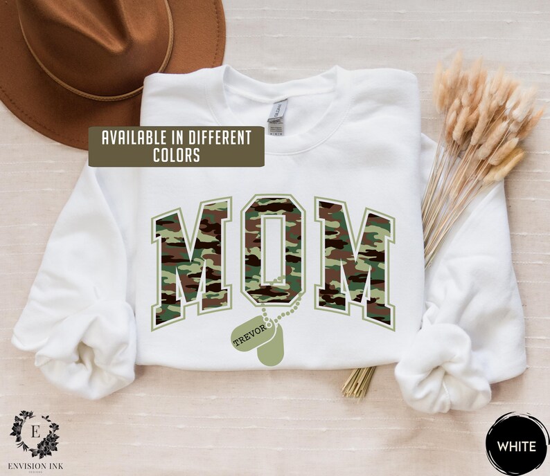 May include: White sweatshirt with a camouflage print of the word "MOM" and a dog tag with the name "TREVOR" on it. The text "AVAILABLE IN DIFFERENT COLORS" is at the top of the image. The word "WHITE" is at the bottom of the image.