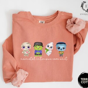 May include: A terracotta orange sweatshirt with a graphic of four cute cartoon monsters. The text "neonatal intensive care unit" is printed below the monsters.