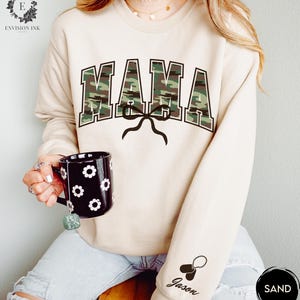 K&ouml;nnte beinhalten: Ein beiger Pullover mit einem Tarnmuster-Druck "MAMA" und einer schwarzen Schleife. Der Pullover hat ein schwarzes Hundemarken mit dem Namen "Jason" am &Auml;rmel.