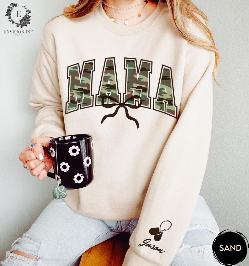 K&ouml;nnte beinhalten: Ein beiger Pullover mit einem Tarnmuster-Druck "MAMA" und einer schwarzen Schleife. Der Pullover hat ein schwarzes Hundemarken mit dem Namen "Jason" am &Auml;rmel.