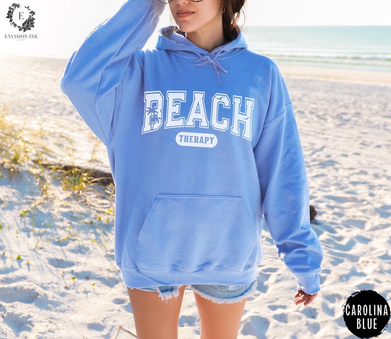 May include: A light blue hoodie with the text "Beach Therapy" in white lettering. The hoodie has a large pocket on the front and a drawstring hood.