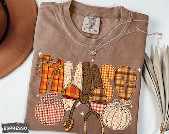 May include: Espresso-colored t-shirt with the word "NURSE" in a fall-themed design. The letters are decorated with plaid, floral, and leopard print patterns. A stethoscope and pumpkins are also featured in the design. A silver necklace is draped across the shirt.