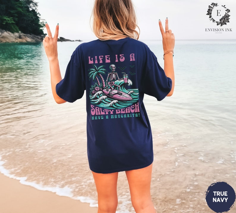 K&ouml;nnte beinhalten: Ein marineblaues T-Shirt mit einem Grafikdruck eines Skeletts auf einem Surfbrett mit dem Text "Life is a Salty Beach Have a Margarita!"