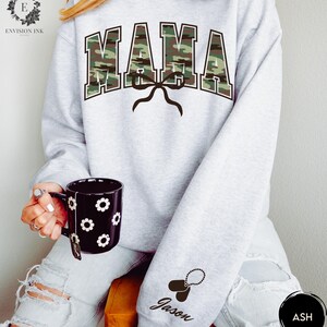 K&ouml;nnte beinhalten: Grauer Pullover mit einem Tarnmuster-Druck "MAMA" und einer schwarzen Schleife. Der Pullover hat ein schwarzes Hundemarken mit dem Namen "Jason" am &Auml;rmel.