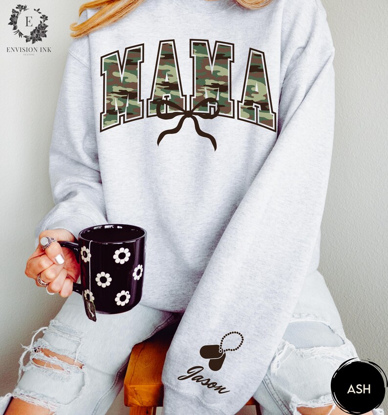 K&ouml;nnte beinhalten: Grauer Pullover mit einem Tarnmuster-Druck "MAMA" und einer schwarzen Schleife. Der Pullover hat ein schwarzes Hundemarken mit dem Namen "Jason" am &Auml;rmel.