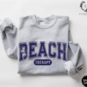 May include: A gray sweatshirt with the text "Beach Therapy" in navy blue letters. The text is in a collegiate font with palm trees and birds on either side.