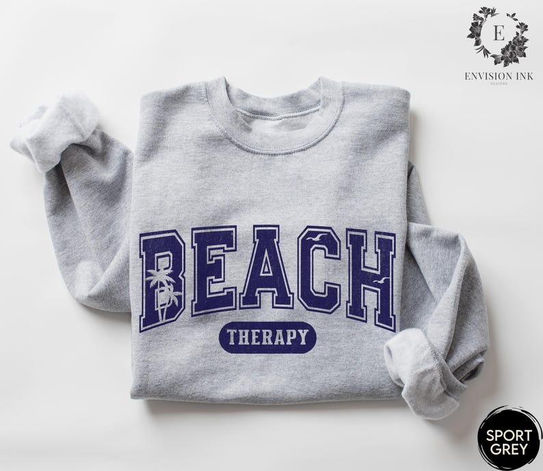May include: A gray sweatshirt with the text "Beach Therapy" in navy blue letters. The text is in a collegiate font with palm trees and birds on either side.
