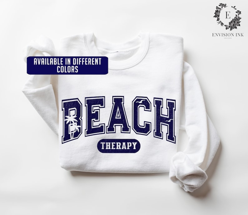 May include: White sweatshirt with a navy blue graphic that says "Beach Therapy" with palm trees and seagulls. The text is in a vintage collegiate font.