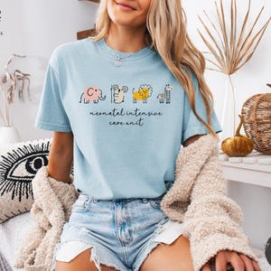 May include: A light blue t-shirt with a graphic of a line of animals, including an elephant, a giraffe, a lion, and a zebra. The text "neonatal intensive care unit" is printed below the animals.