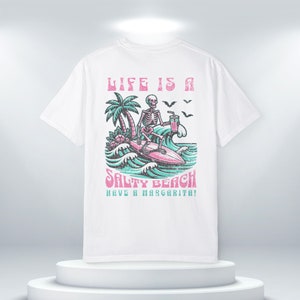 K&ouml;nnte beinhalten: Wei&szlig;es T-Shirt mit einem Grafikdruck eines Skeletts, das mit einer Margarita surft. Der Text "Life is a Salty Beach Have a Margarita!" ist auf dem T-Shirt aufgedruckt.