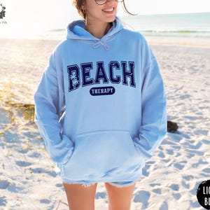 May include: A light blue hoodie with the text "Beach Therapy" in navy blue lettering. The text is in a collegiate font with palm trees on either side.