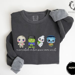 May include: A dark grey sweatshirt featuring a graphic of four cartoon monsters: a mummy, Frankenstein, a ghost, and a zombie. The text "neonatal intensive care unit" is printed below the graphic.