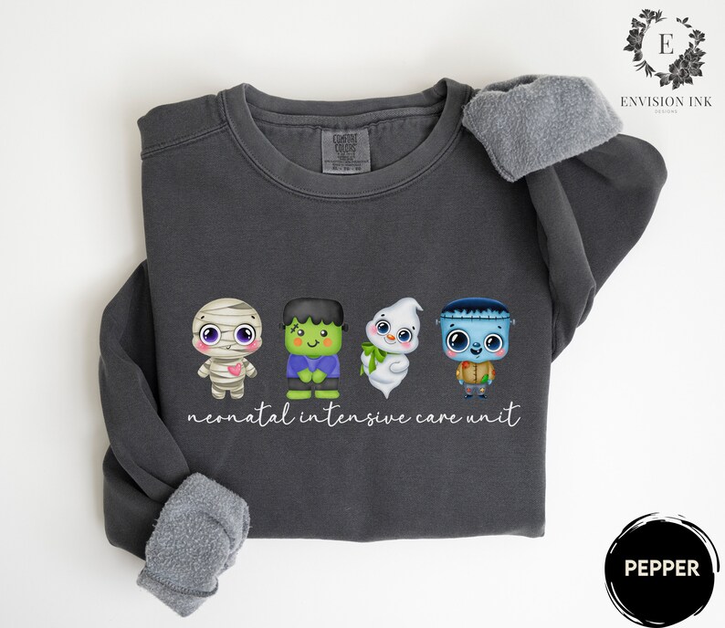 May include: A dark grey sweatshirt featuring a graphic of four cartoon monsters: a mummy, Frankenstein, a ghost, and a zombie. The text "neonatal intensive care unit" is printed below the graphic.