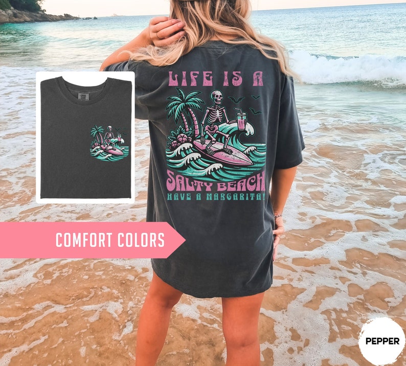 K&ouml;nnte beinhalten: Ein dunkelgraues T-Shirt mit einem Grafikdruck eines Skeletts, das auf einer Welle mit einer Margarita surft. Der Text auf dem Shirt lautet "Life is a Salty Beach Have a Margarita!"