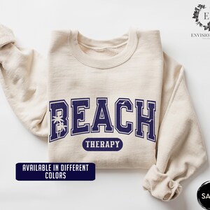 May include: A beige sweatshirt with the text "Beach Therapy" in navy blue lettering. The text is in a collegiate font with palm trees and seagulls on either side. The sweatshirt is available in different colors.
