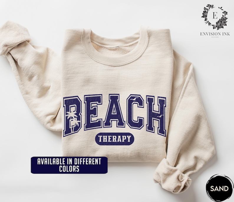 May include: A beige sweatshirt with the text "Beach Therapy" in navy blue lettering. The text is in a collegiate font with palm trees and seagulls on either side. The sweatshirt is available in different colors.