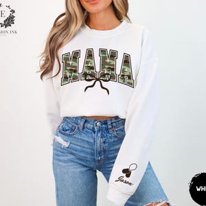 K&ouml;nnte beinhalten: Wei&szlig;er Pullover mit dem Wort "MAMA" in Camouflage-Druck. Der Pullover hat eine Schleife und ein Hundemarken mit dem Namen "Jason" am &Auml;rmel.