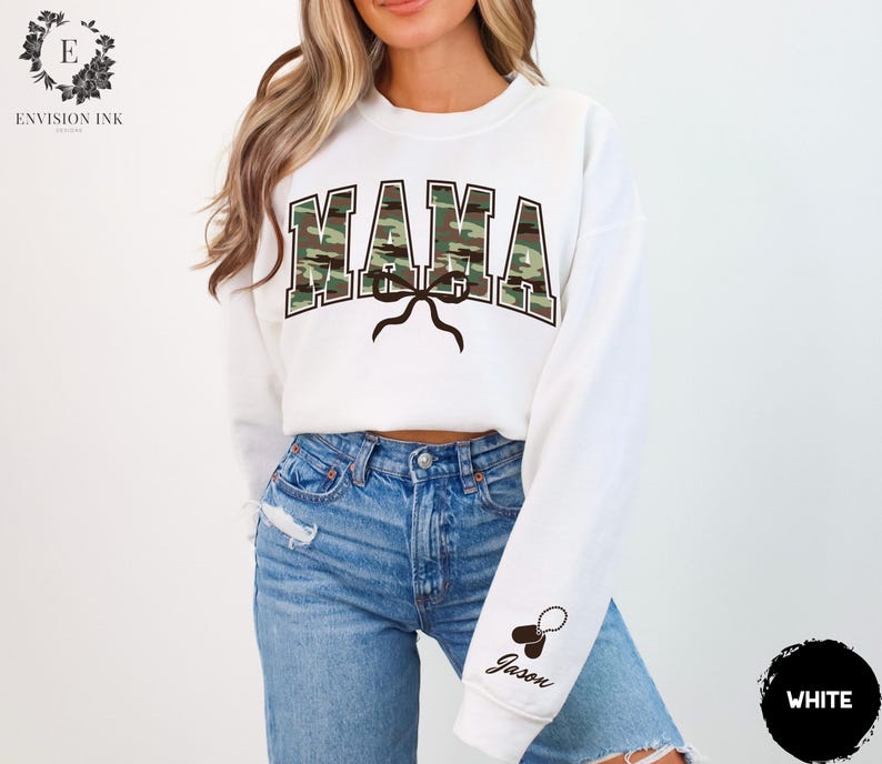 K&ouml;nnte beinhalten: Wei&szlig;er Pullover mit dem Wort "MAMA" in Camouflage-Druck. Der Pullover hat eine Schleife und ein Hundemarken mit dem Namen "Jason" am &Auml;rmel.