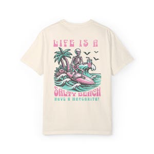 K&ouml;nnte beinhalten: Ein hellbeiges T-Shirt mit einem Grafikdruck eines Skeletts, das surft und ein Getr&auml;nk h&auml;lt. Der Text "Life is a Salty Beach Have a Margarita!" ist unter dem Grafikdruck aufgedruckt.