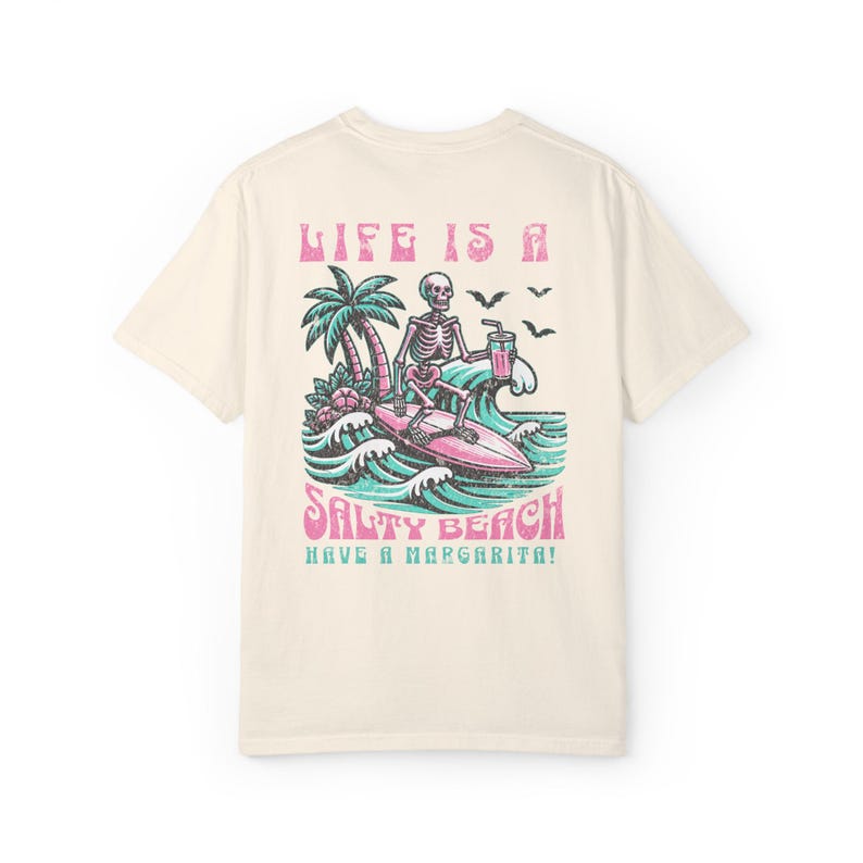 K&ouml;nnte beinhalten: Ein hellbeiges T-Shirt mit einem Grafikdruck eines Skeletts, das surft und ein Getr&auml;nk h&auml;lt. Der Text "Life is a Salty Beach Have a Margarita!" ist unter dem Grafikdruck aufgedruckt.