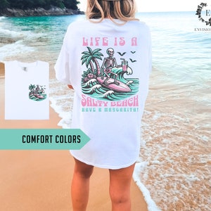 K&ouml;nnte beinhalten: Wei&szlig;es T-Shirt mit einem Grafikdruck eines Skeletts, das auf einer Welle surft, mit dem Text "Life is a Salty Beach Have a Margarita!"