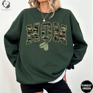 May include: Forest green sweatshirt with a camouflage print of the word "MOM" and a dog tag with the name "TREVOR" printed on it.