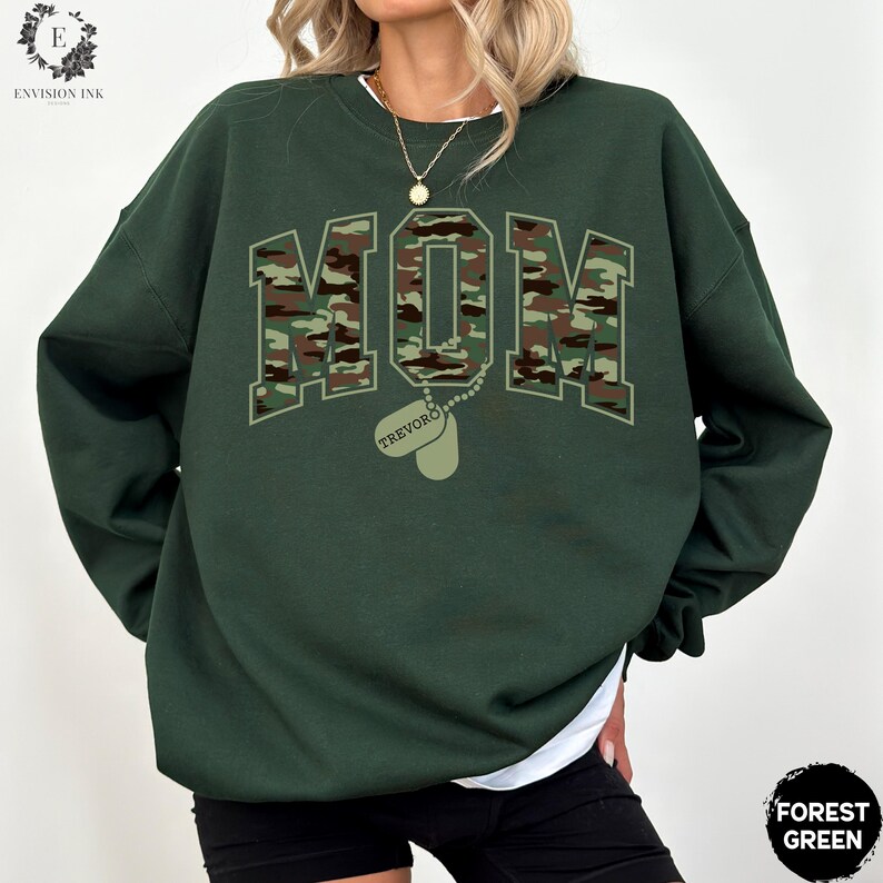 May include: Forest green sweatshirt with a camouflage print of the word "MOM" and a dog tag with the name "TREVOR" printed on it.
