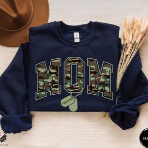 May include: Navy blue sweatshirt with a camouflage print of the word "MOM" and a dog tag with the name "TREVOR" printed on it.