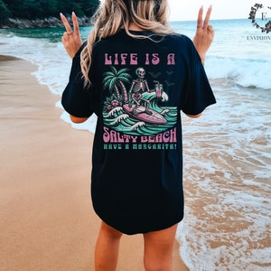 K&ouml;nnte beinhalten: Schwarzes T-Shirt mit einem Grafikdruck eines Skeletts, das mit einer Margarita surft. Der Text auf dem Shirt lautet "Life is a Salty Beach Have a Margarita!"