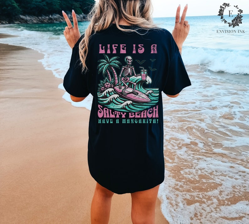 K&ouml;nnte beinhalten: Schwarzes T-Shirt mit einem Grafikdruck eines Skeletts, das mit einer Margarita surft. Der Text auf dem Shirt lautet "Life is a Salty Beach Have a Margarita!"