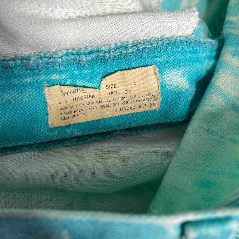 Op de afbeelding: Een blauw en wit tie-dye Wrangler jeans label met het stylenummer NA687AA, maat 5, lengte 32 en wasinstructies. Het label zegt "Machinewas met soortgelijke kleuren. Alleen niet-chloor bleekmiddel gebruiken indien nodig. Drogen in de droger, direct verwijderen. Gemaakt in de VS."