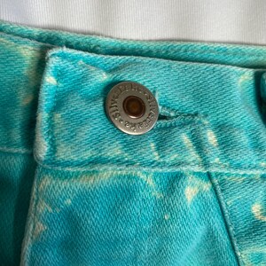Op de afbeelding: Close-up van een paar blauwe denim jeans met een wit tie-dye patroon. De jeans heeft een metalen knoop met de tekst "Stella Jeans" erop.
