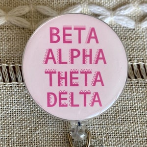 May include: A white round badge with a silver metal frame and a clear plastic retractable badge holder. The badge has pink text that reads "Beta Alpha Theta Delta".