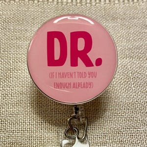 May include: Pink round badge holder with a silver clip and a pink circle with the text "DR. (IF I HAVEN'T TOLD YOU ENOUGH ALREADY)"