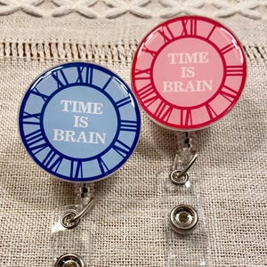 Może przedstawiać: Dwa okrągłe zwijacze do identyfikatorów z motywami tarcz zegarowych. Jeden jest niebieski z białym tekstem „TIME IS BRAIN”. Drugi jest różowy z czerwonym tekstem, który mówi to samo. Oba mają przezroczyste, plastikowe sznurki i metalowe klipsy.