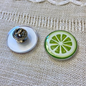 May include: Two round enamel pins. One pin features a lime slice design with green and white colors. The other pin is white with a silver pin back. The pins are on a beige textured surface.