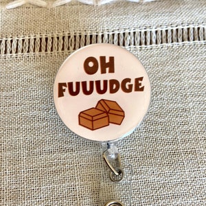 May include: A white round badge holder with a silver metal clip. The badge has a pink background with the text "OH FUUUDGE" in brown letters and a graphic of two brown chocolate squares.