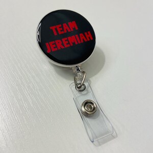 May include: A black and silver retractable badge reel with the text "TEAM JEREMIAH" in red. The badge reel has a clear plastic strap with a metal snap closure. The reel is round and has a glossy finish.